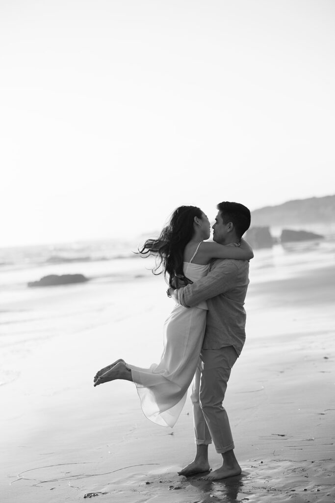 Black and white engagement photo of man twirling woman on Crystal Cove beach.