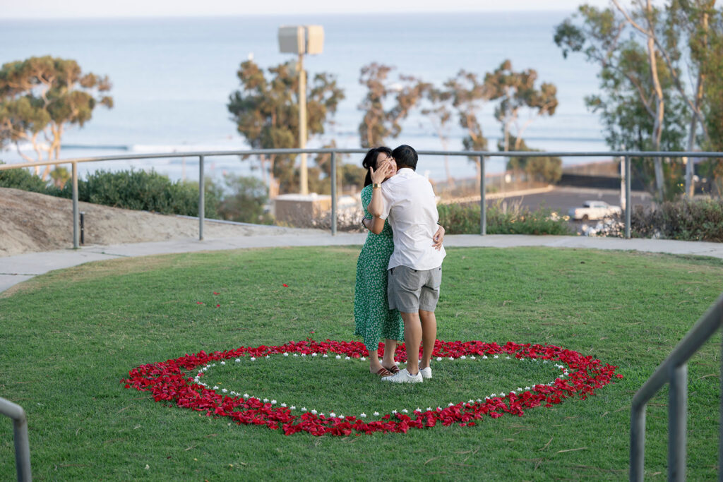 Man and woman hugging after surprise proposal at Dana Point surrounded by roses petals and candles arranged in large heart.