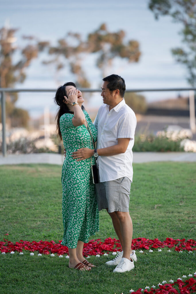 Woman wipes tear away as she smiles after proposal at Dana Point surrounded by rose petals and candles.