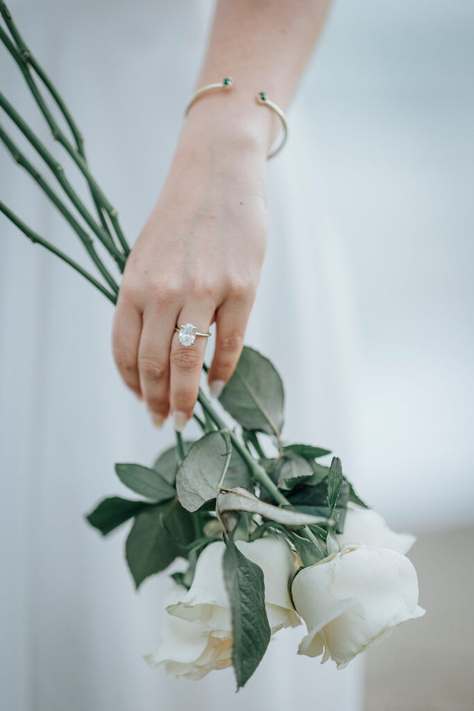 Close up photo of woman's engagement ring as she holds bouquet of white roses on the beach after her proposal in Orange County. 