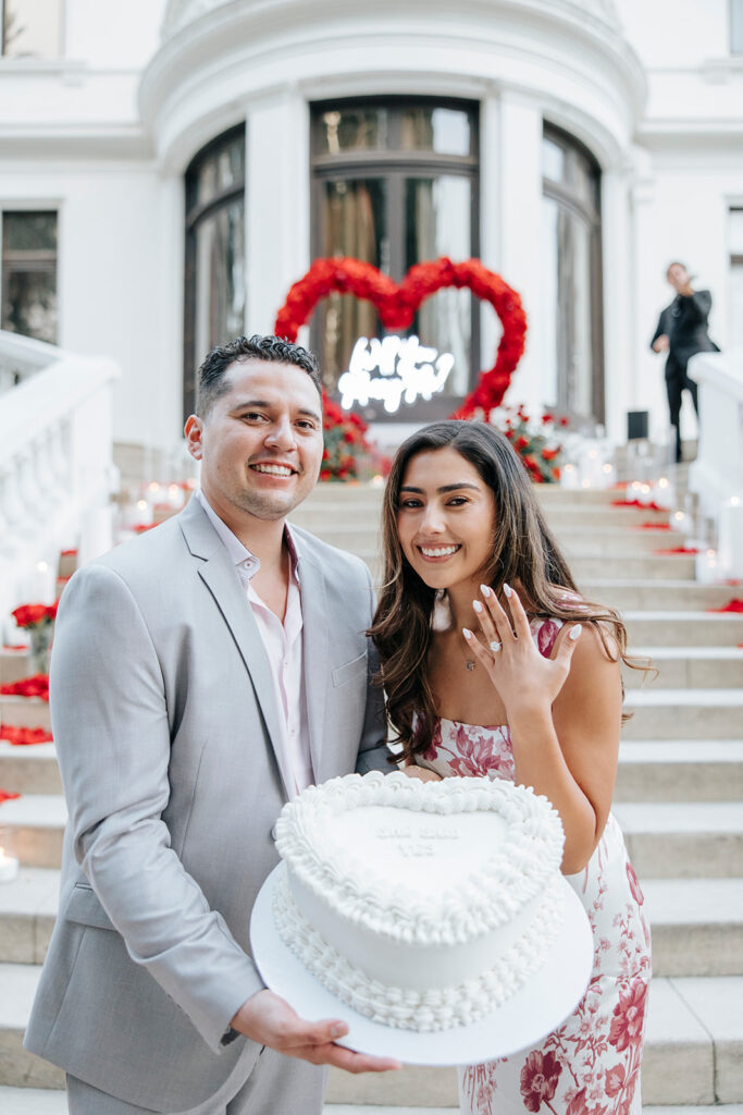 Couple stands in front of Pasadena Museum of History with white vintage heart cake as woman shows off engagement ring to proposal photographer.