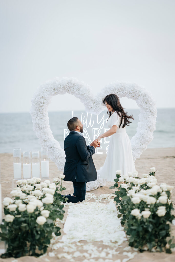 Man proposing on Orange County beach in front of large heart-shaped floral installment, candles, and aisle of white roses.