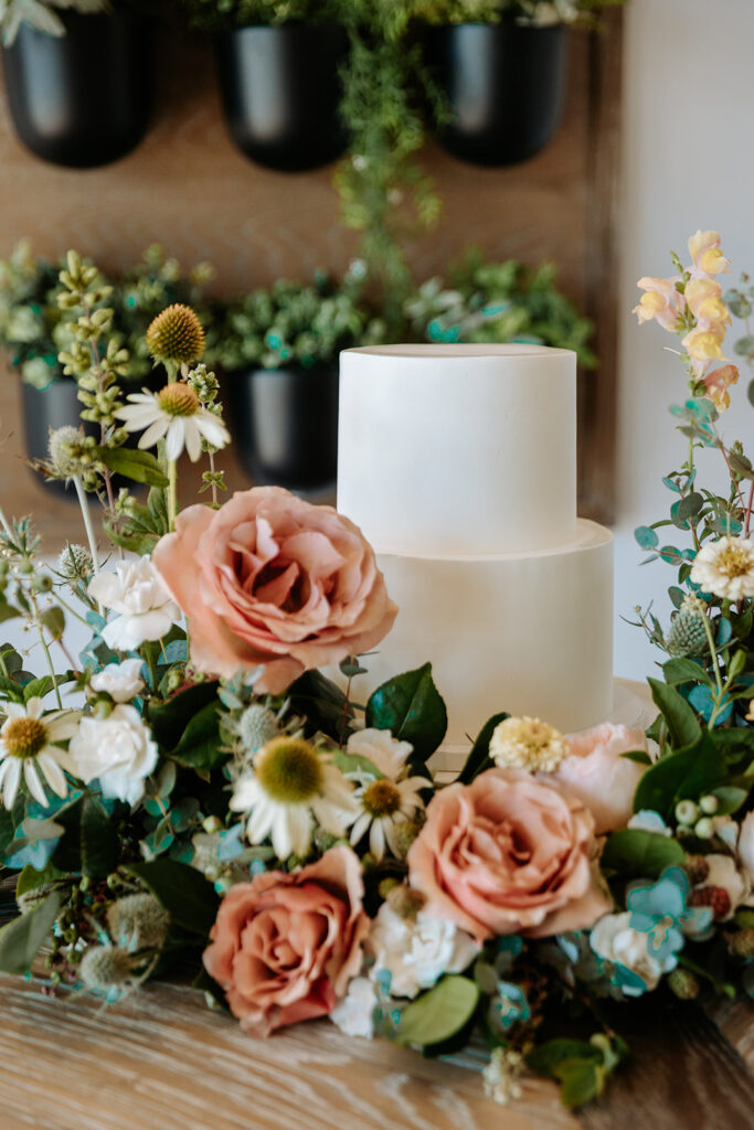 simple white 2 tier wedding cake with cake garden surrounding it and hanging plant display behind 