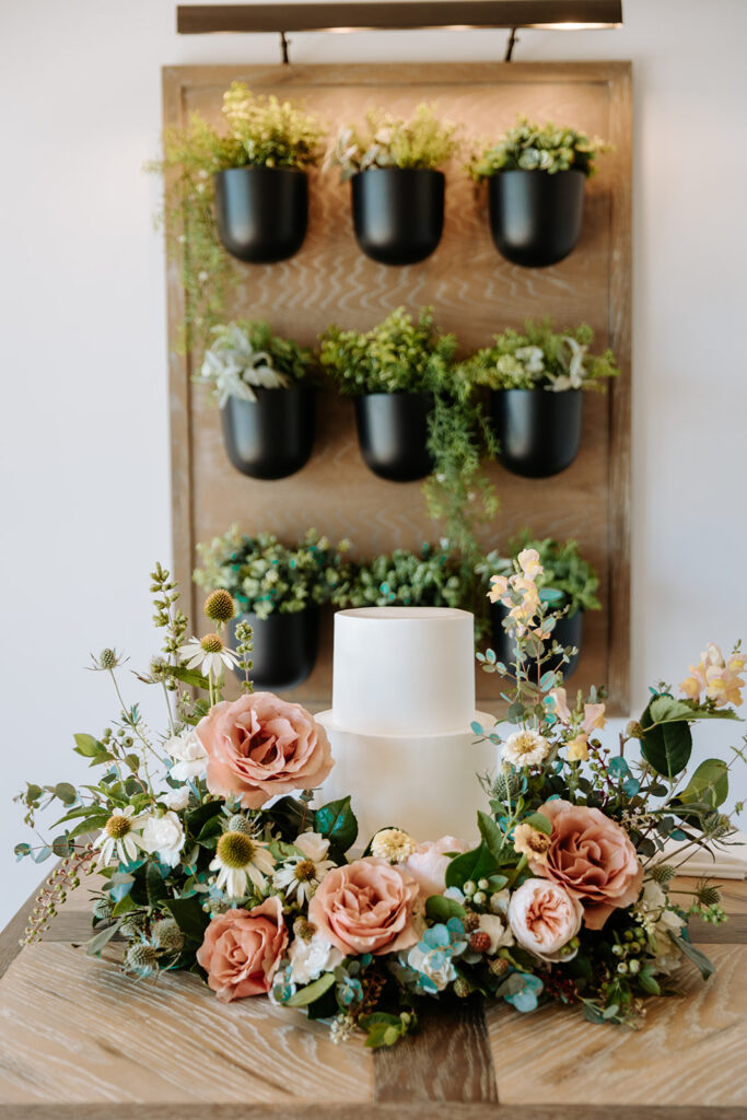 simple white 2 tier wedding cake with cake garden surrounding it and hanging plant display behind 