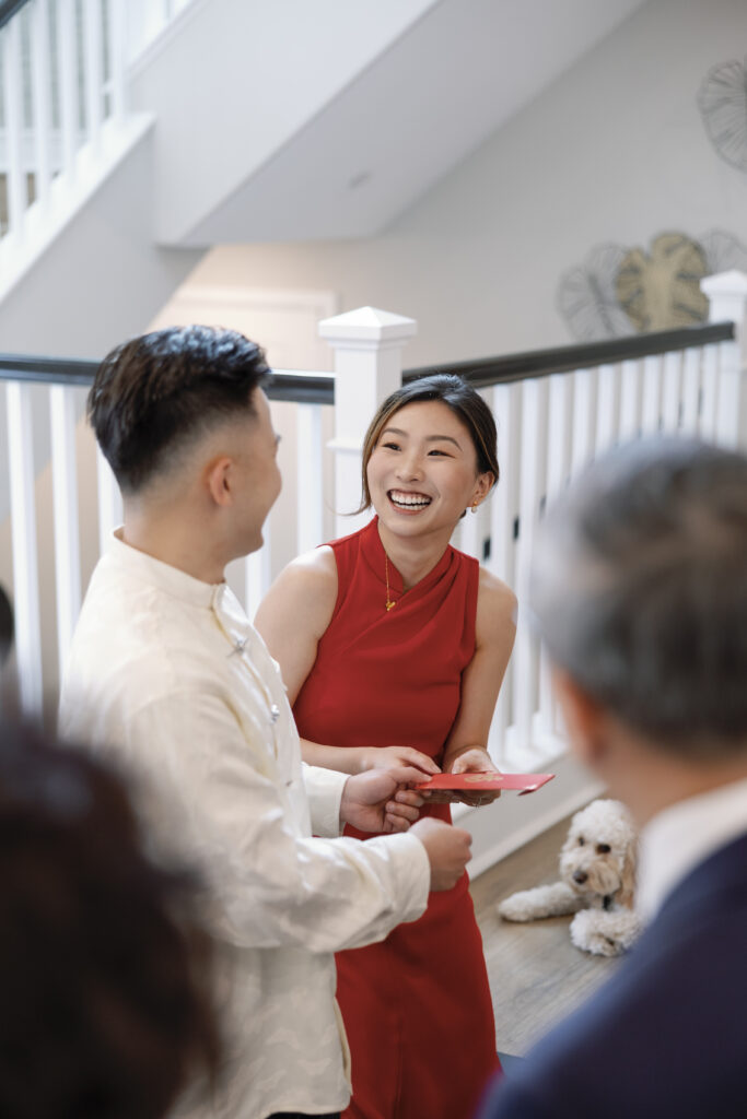 A bride in a red cheongsam sharing a joyful moment with her groom during a tea ceremony, with family (and a cute pup!) nearby