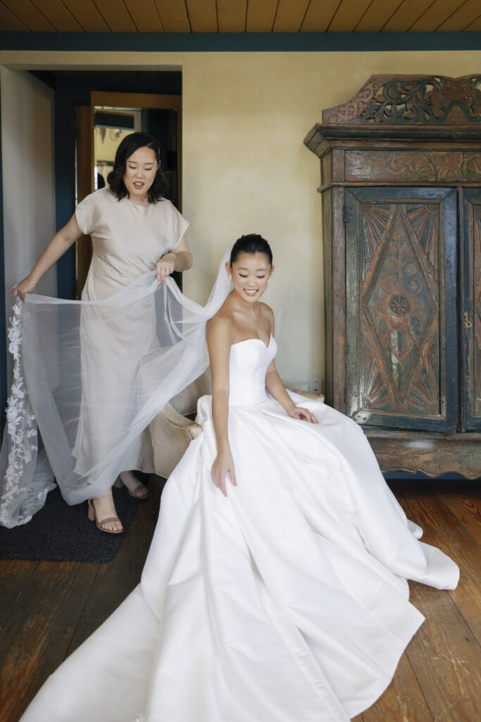 a bride sitting down while her bridesmaid adjusts and spreads out her veil