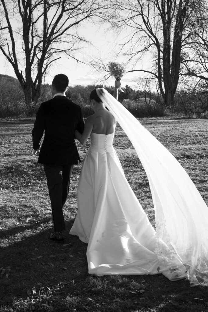 black and white photo of bride and groom walking away outside. bride has bouquet held in the air in celebration and vieil flowing and trailing behind the bride