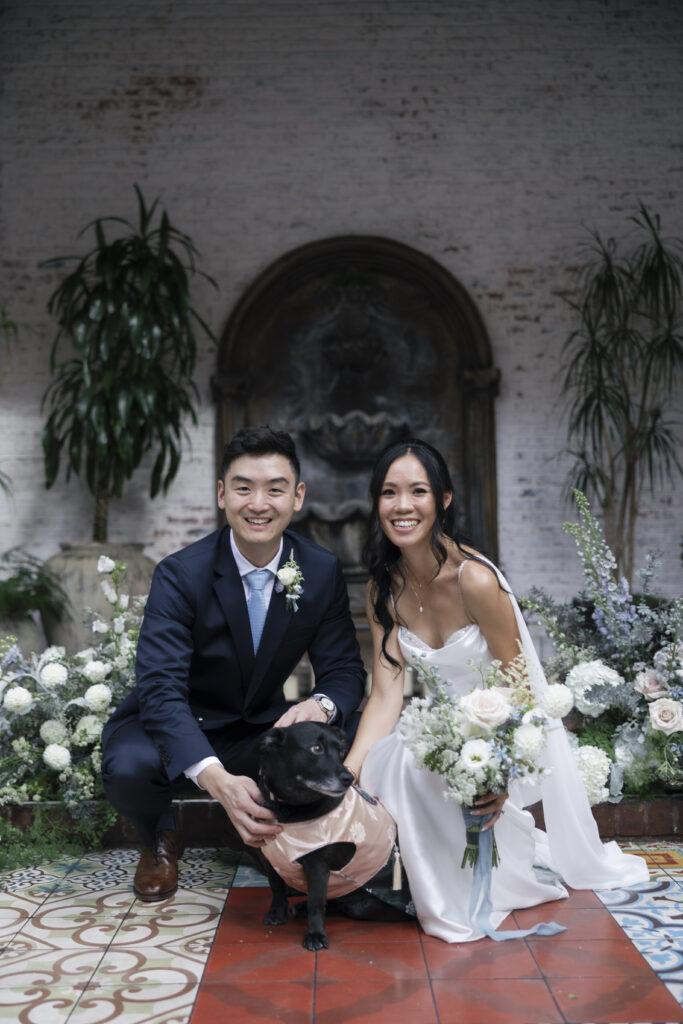 bride and groom posing with their dog. the dog is dressed in a cute pink outfit