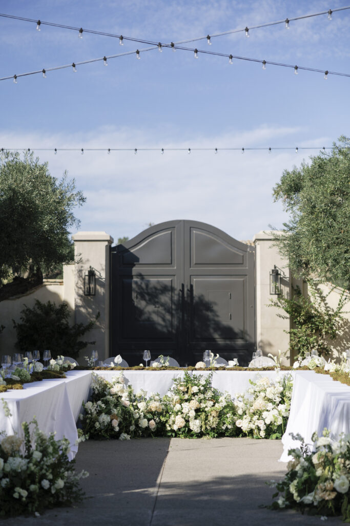 A courtyard reception setup with elegant white floral-lined tables, soft string lights, and a grand gate in the background.