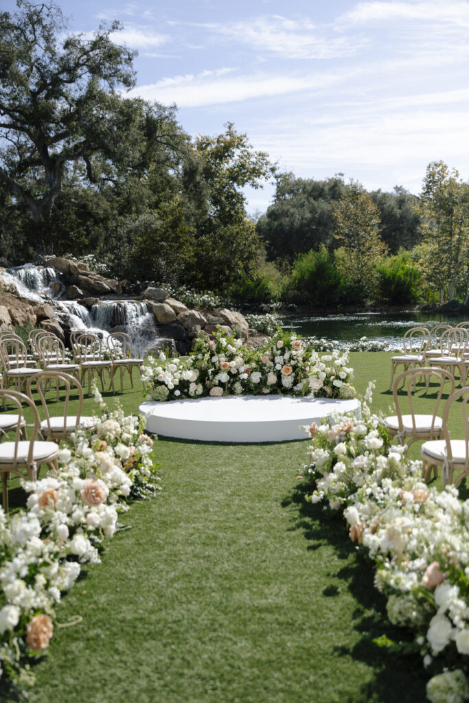 A serene outdoor ceremony with lush white florals lining the aisle and circular ceremony platform. set against a backdrop of greenery and a peaceful waterfall.