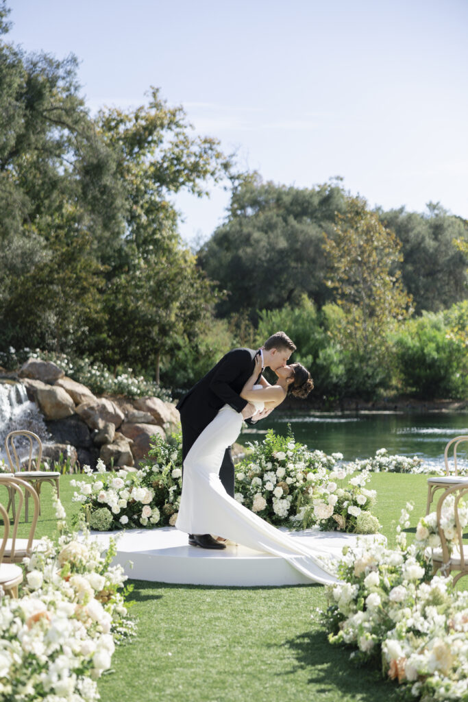 A romantic couple's kiss on a white circular platform, surrounded by soft white floral arrangements and a lush, greenery and river backdrop.