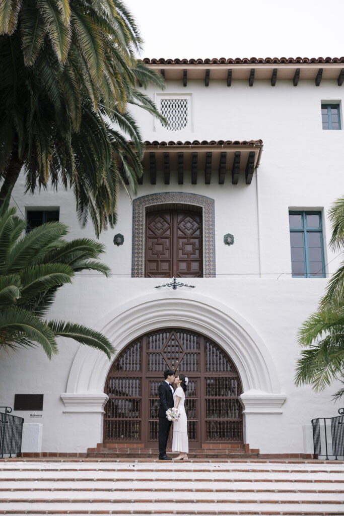 Elopement at Santa Barbara Courthouse with a couple kissing in front of the iconic arched wooden door, framed by Spanish-style architecture and lush palm trees.