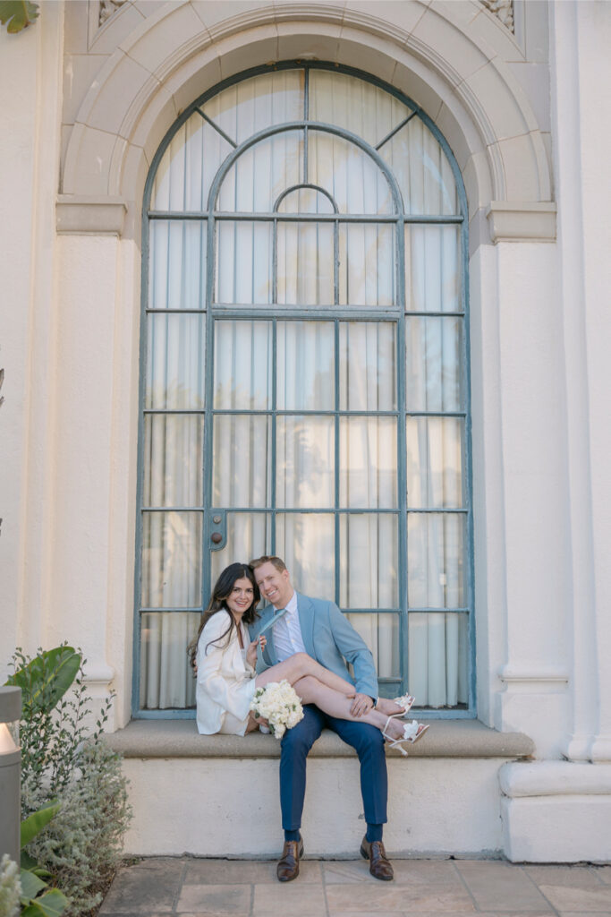 A joyful couple poses sitting on a windowsill at the Beverly Hills Courthouse, celebrating their city hall elopement in Orange County.