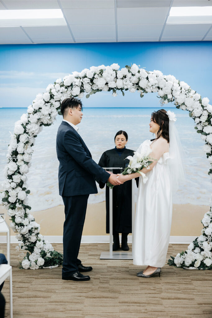 Bride and groom exchange vows during city hall elopement in Orange County, with white floral arch around them. 