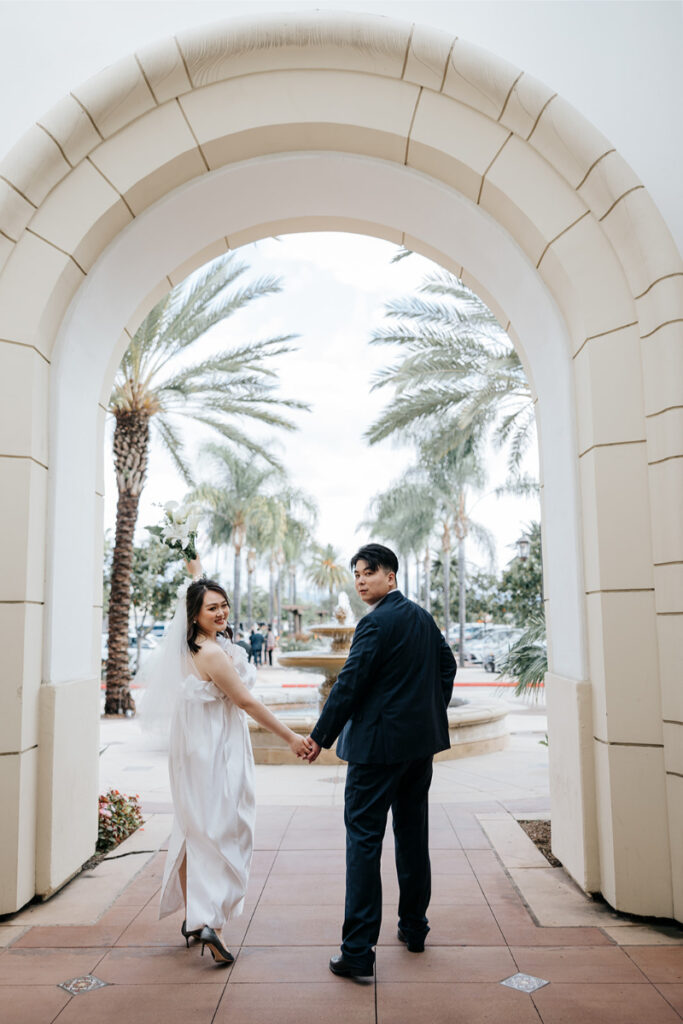 Bride and groom holding hands as they walk away from photographer and towards fountain at Laguna Hills Civic Center. 