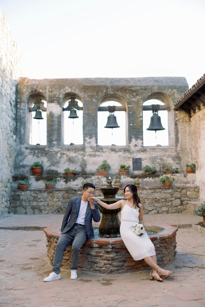 A couple posing near a beautiful fountain with historic bell arches in the background at an Orange County wedding venue.