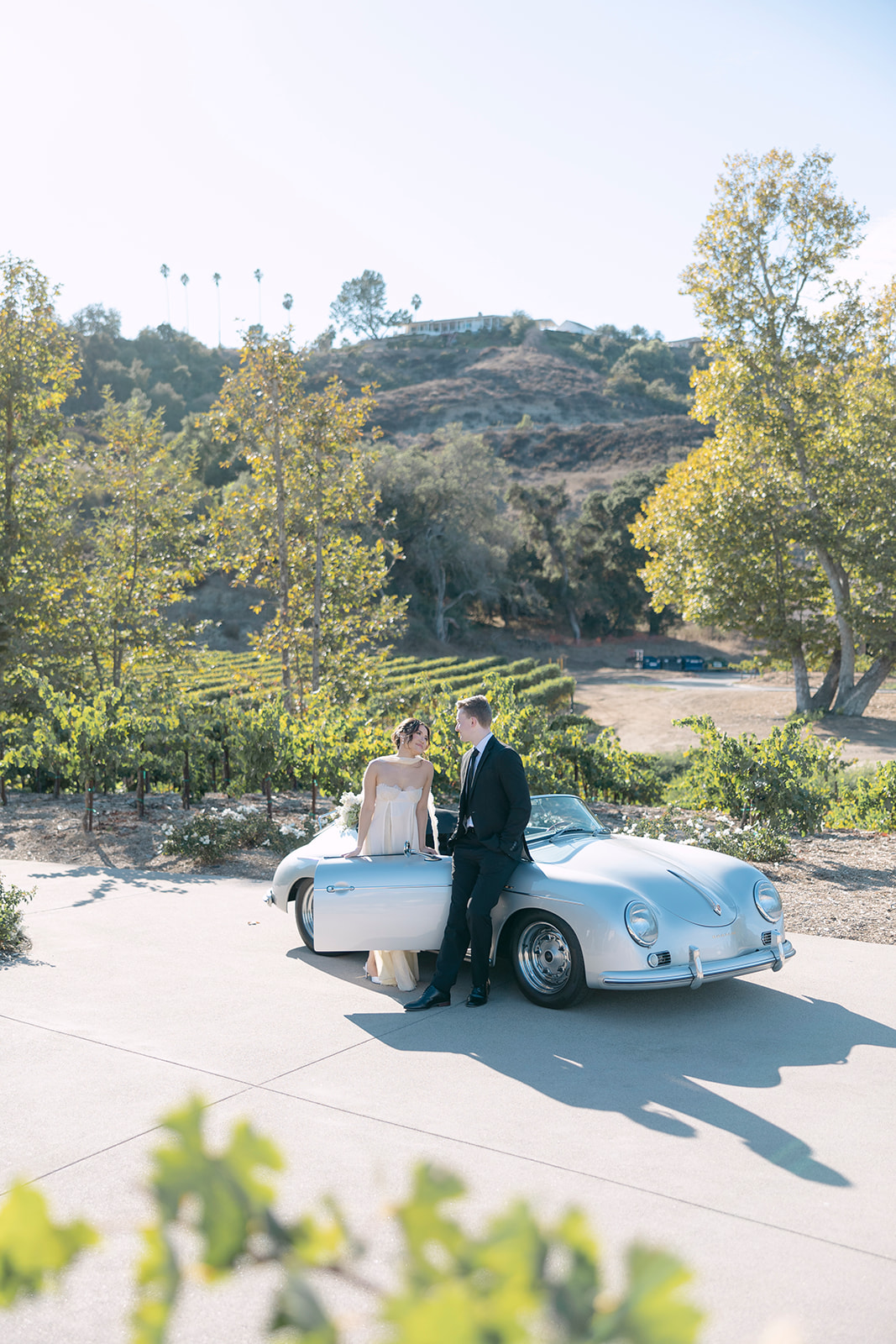 A couple standing beside a classic car in a vineyard setting, celebrating their engagement.