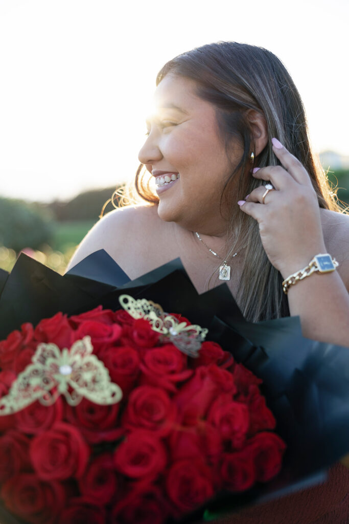 A close-up of a bride-to-be smiling, holding a bouquet of red roses with gold butterfly details.