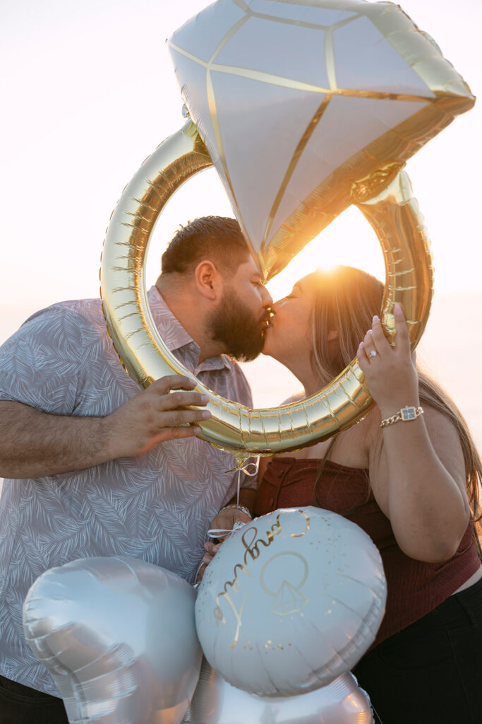 A couple celebrating being engaged in Orange County, sharing a kiss through a golden engagement ring-shaped balloon with the sun in the background.