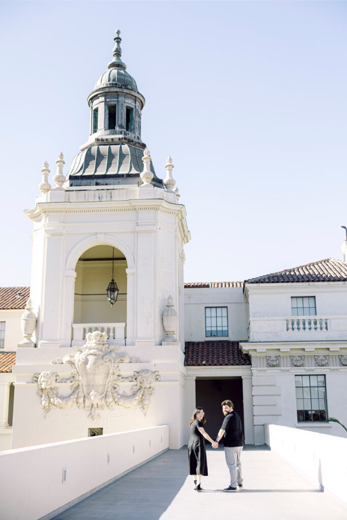 A couple walks hand-in-hand across the Pasadena City Hall bridge, framed by its iconic dome and intricate architectural details.