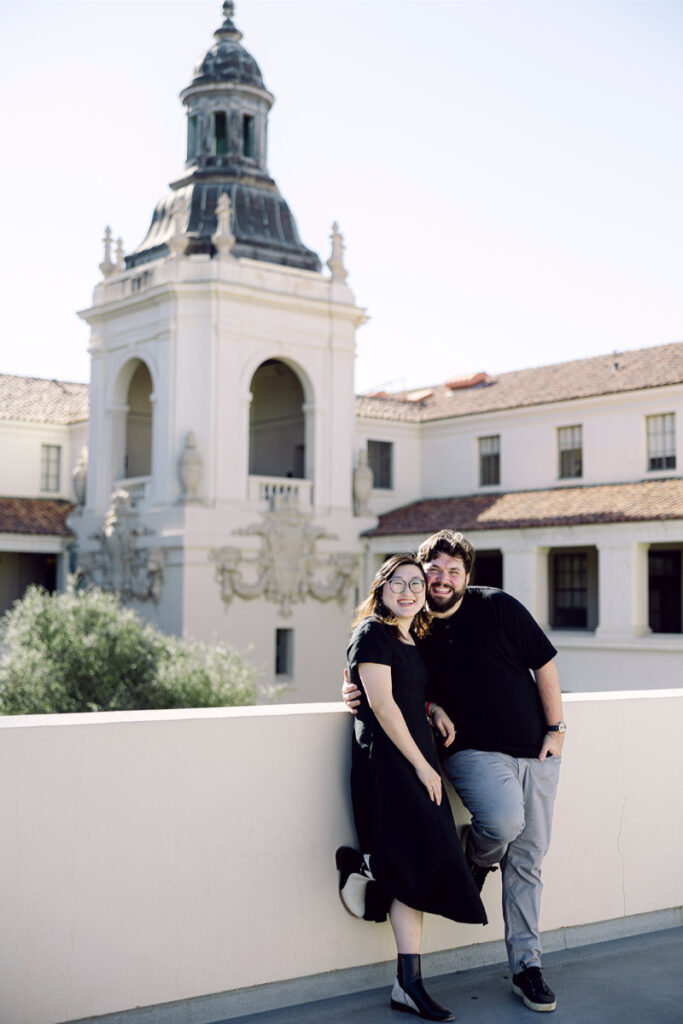 The couple poses together, leaning against a railing with the beautiful Pasadena City Hall as the backdrop.