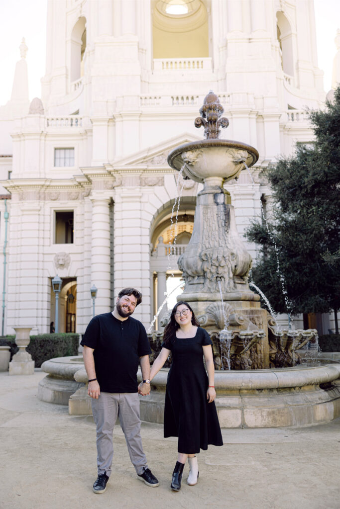 The couple smiles in front of the grand fountain at Pasadena City Hall, celebrating their special day.