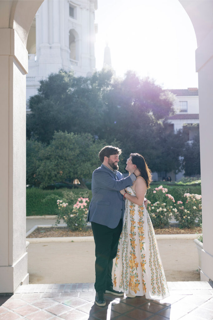 Bathed in sunlight, the bride and groom embrace under an arch of Pasadena City Hall’s with flower garden behind them. 