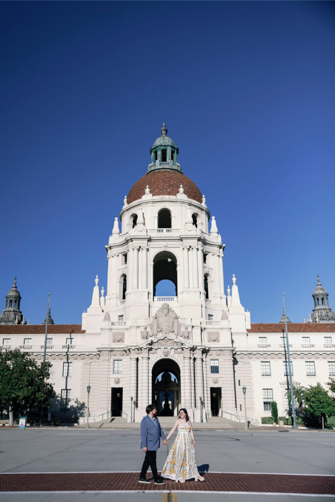 A wide-angle view of the couple walking in front of the stunning Pasadena City Hall dome.