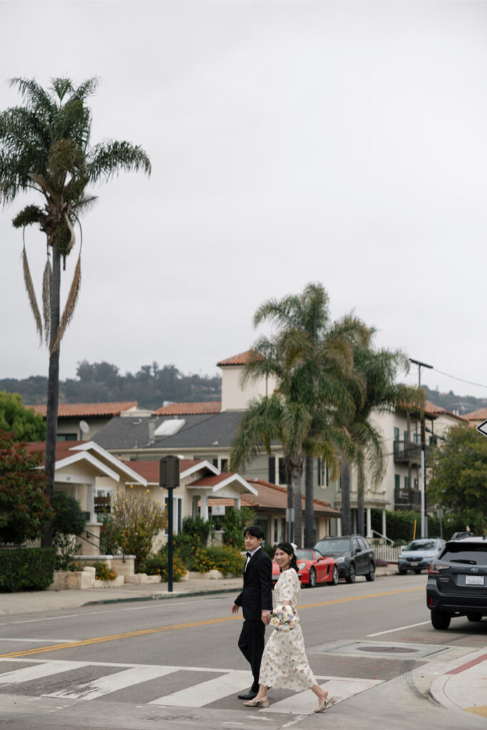 Bride and groom hold hands as they cross street together after elopement. 